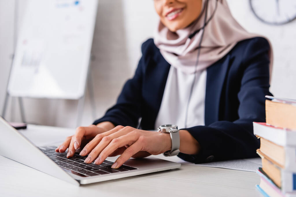 przycięty widok uśmiechniętego arabskiego tłumacz wpisując na laptopie w pobliżu słowników języków obcych, rozmyte tło - Zdjęcie, obraz