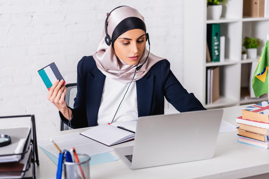 αραβικός διερμηνέας στα ακουστικά που κρατά τον ψηφιακό μεταφραστή ενώ εργάζεται στο lap-top κοντά στα λεξικά, θολό προσκήνιο - Φωτογραφία, εικόνα