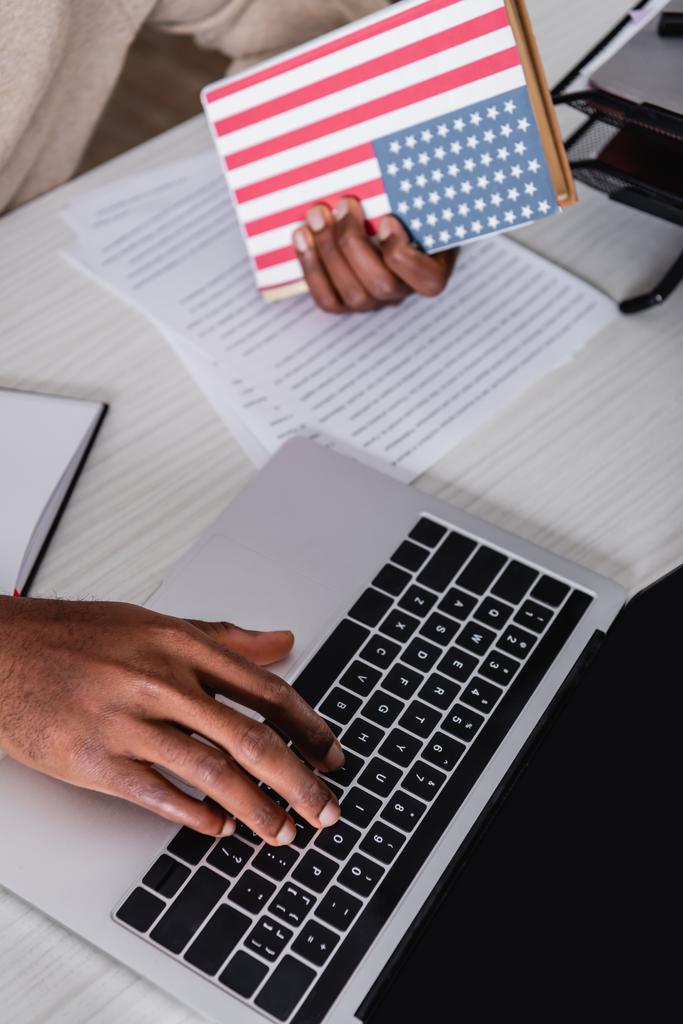 Ausgeschnittene Ansicht eines afrikanisch-amerikanischen Übersetzers, der auf seinem Laptop tippt, während er ein Wörterbuch mit US-Flagge auf dem Cover hält - Foto, Bild