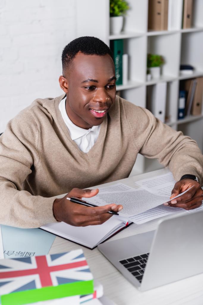 Traducteur afro-américain souriant travaillant avec des documents près d'un ordinateur portable et des dictionnaires sur le premier plan flou - Photo, image