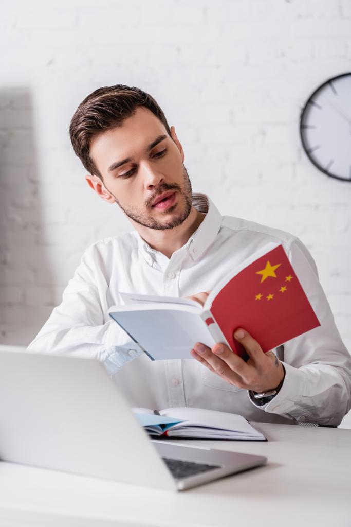 jovem tradutor segurando dicionário chinês perto notebook e laptop em primeiro plano borrado - Foto, Imagem