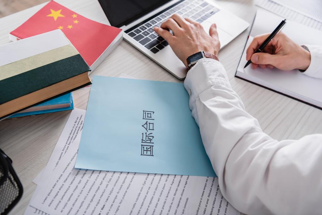частковий вид перекладача, що пише в блокноті та друкує на ноутбуці біля паперу з китайськими ієрогліфами. Переклад: "Міжнародний контракт
" - Фото, зображення
