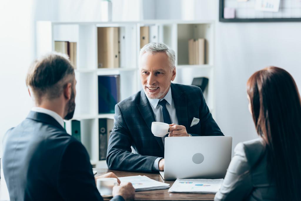 investidor sorridente com xícara de café olhando para os empresários em primeiro plano borrado perto de papéis e laptop  - Foto, Imagem