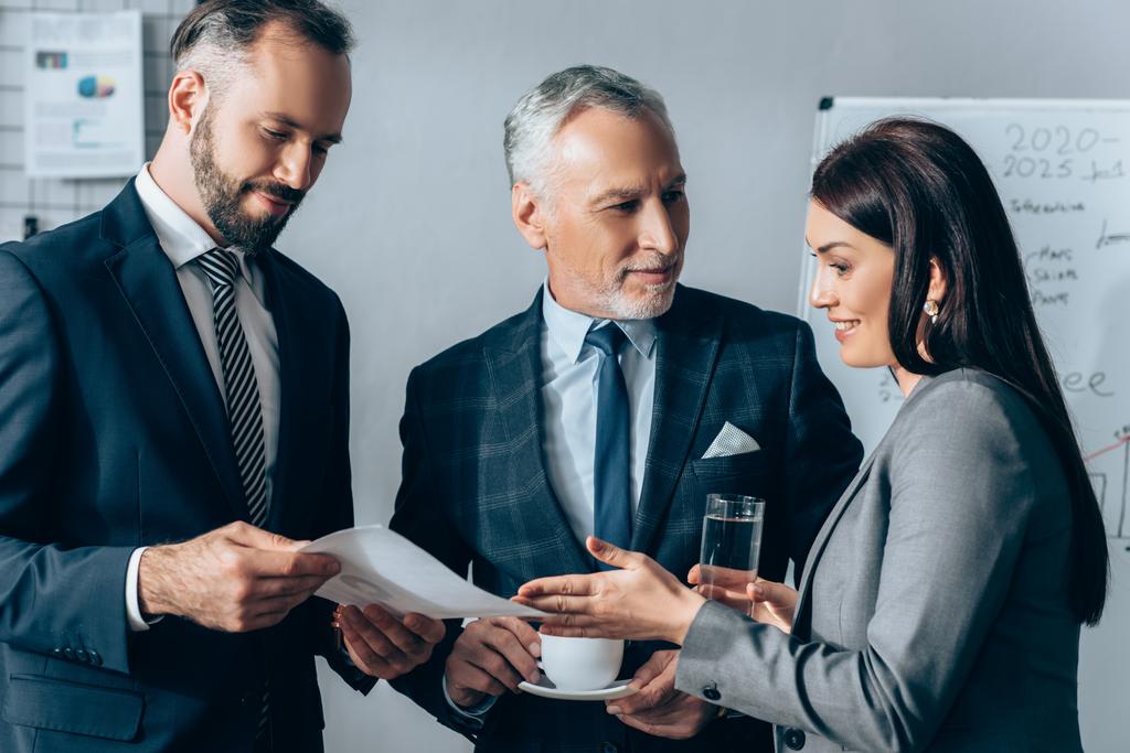 Smiling businesswoman with water pointing at paper near investor with cup and colleague with document in office  - Photo, Image
