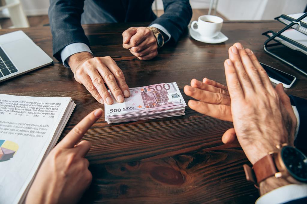 Przycięty widok przedsiębiorców pokazujących gest zatrzymania i wskazujących na banknoty euro w pobliżu kolegi oraz papiery na niewyraźnej nowej wiedzy  - Zdjęcie, obraz