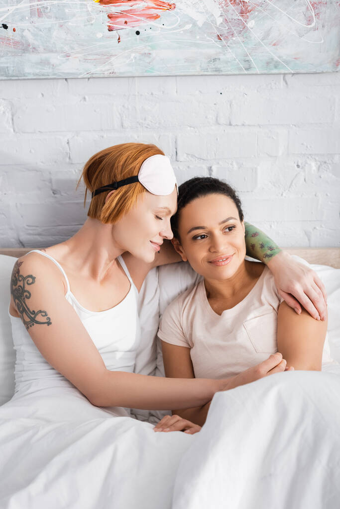 tattooed lesbian woman in eyemask on forehead hugging african american girlfriend in bed - Photo, Image