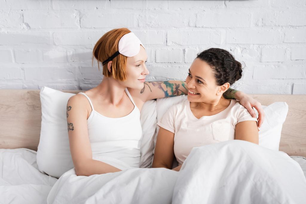 щаслива міжрасова лесбіянка пара дивиться один на одного, сидячи в ліжку
 - Фото, зображення
