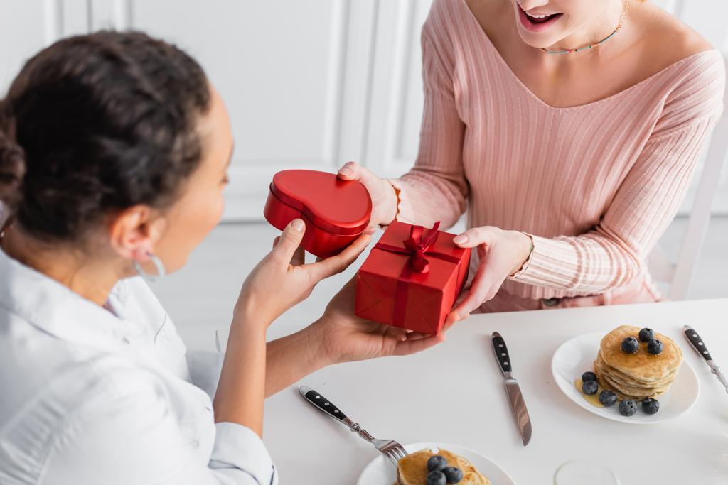 pareja lésbica interracial regalándose regalos durante el desayuno en el día de San Valentín, borrosa en primer plano - Foto, Imagen