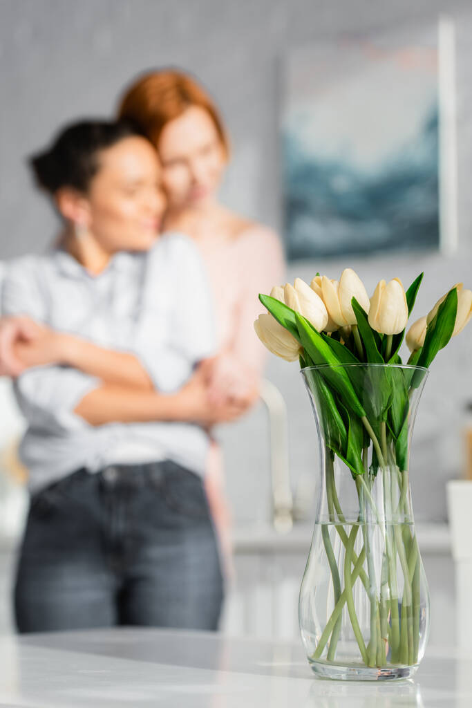 selettiva messa a fuoco di tulipani in vaso vicino interrazziale lesbica coppia abbracci su sfondo sfocato - Foto, immagini