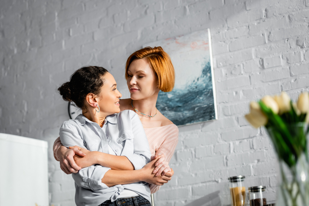 feliz interracial lesbianas pareja abrazando y mirando uno al otro en cocina en borrosa primer plano - Foto, imagen
