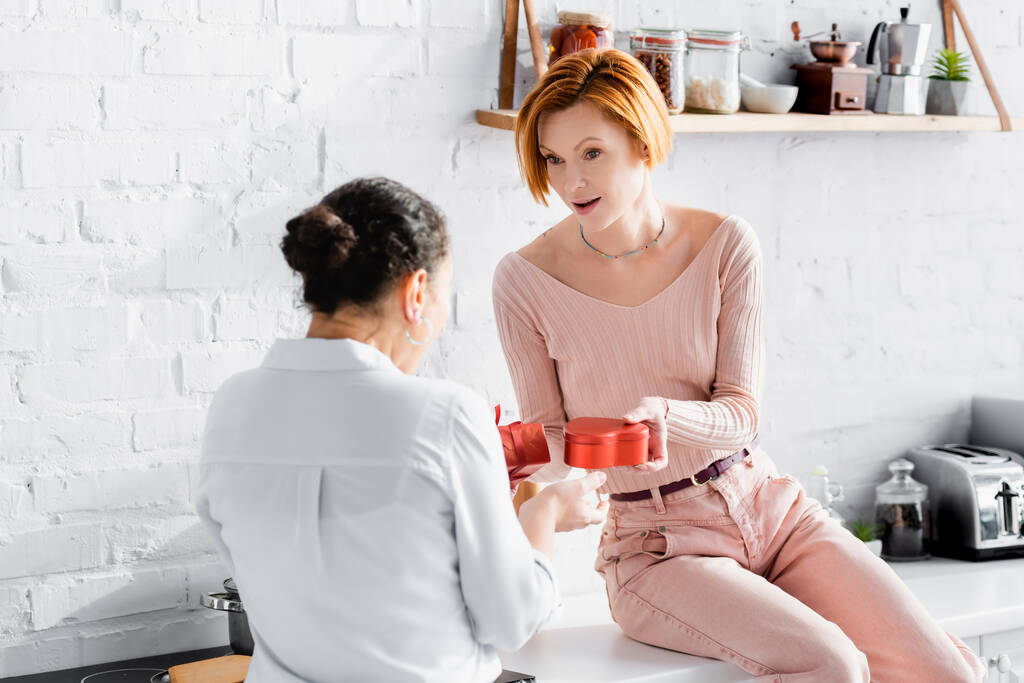 redhead woman presenting gifts to african american girlfriend while sitting on kitchen counter - Photo, Image