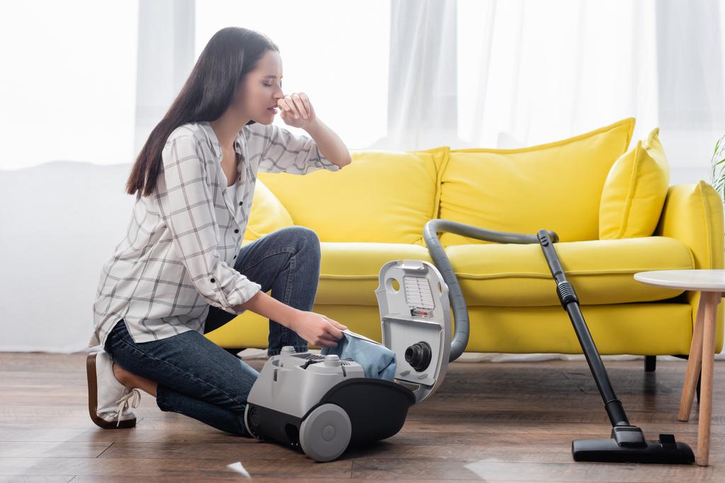 young woman suffering from allergy while holding dust bag near vacuum cleaner - Photo, Image