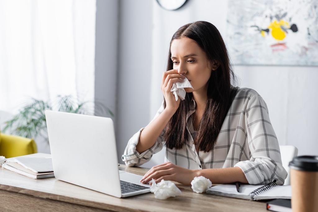 Alerjik bir kadın, ön planda laptopta çalışırken kağıt peçeteyle burnunu siliyor. - Fotoğraf, Görsel