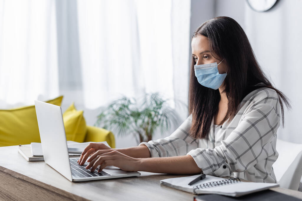 фрилансер в медицинской маске печатает на ноутбуке рядом с ноутбуками дома - Фото, изображение