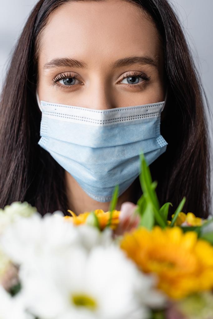 πορτρέτο της αλλεργικής γυναίκας σε ιατρική μάσκα κοιτάζοντας κάμερα κοντά σε λουλούδια, θολή πρώτο πλάνο - Φωτογραφία, εικόνα