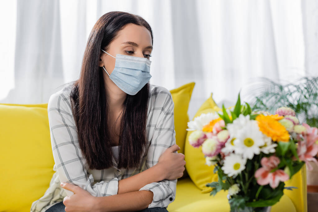 Üzgün, alerjik, maskeli bir kadın. Çiçeklerin yanında oturuyor. - Fotoğraf, Görsel
