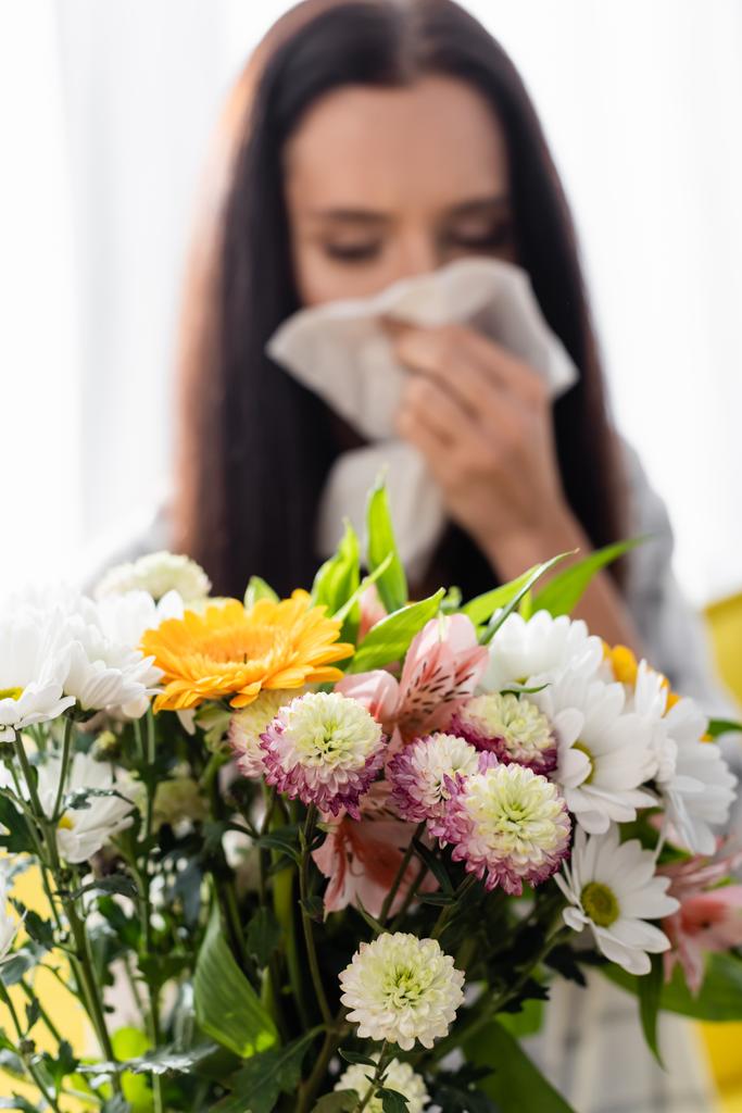 selektivní zaměření kytice v blízkosti alergické ženy otírání nosu s papírovým ubrouskem na rozmazaném pozadí - Fotografie, Obrázek