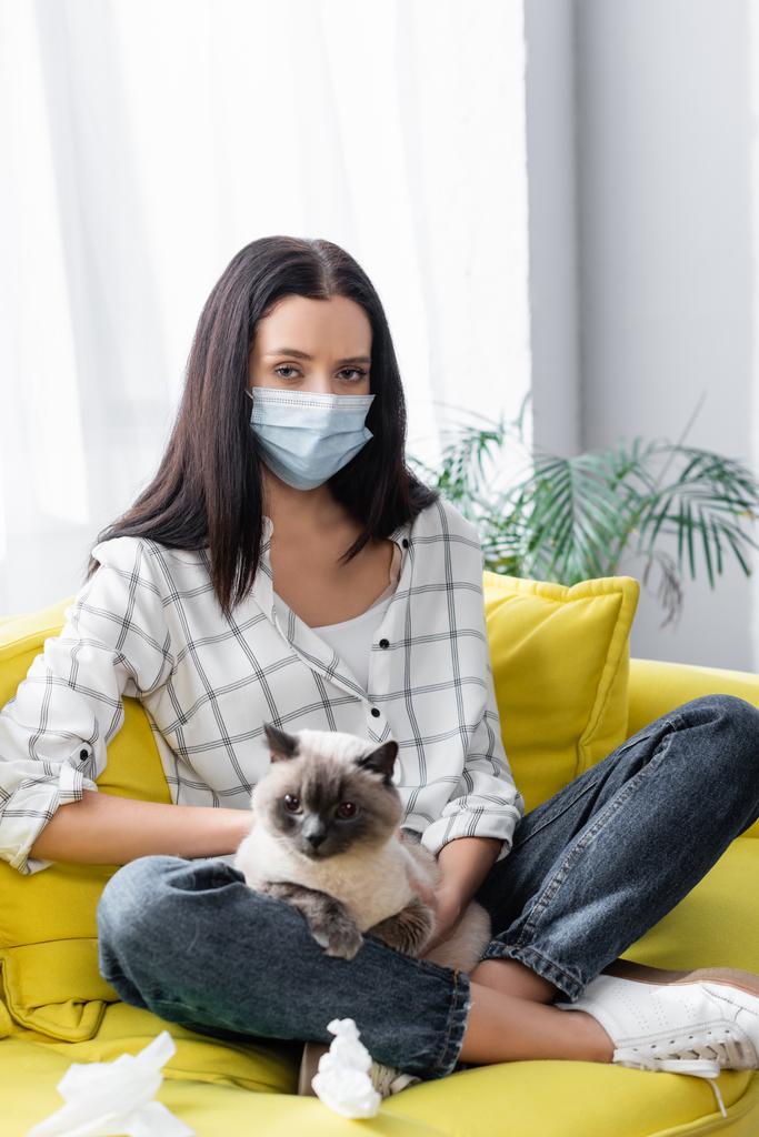 uczulona kobieta w masce medycznej patrząc na aparat podczas siedzenia na kanapie z kotem - Zdjęcie, obraz