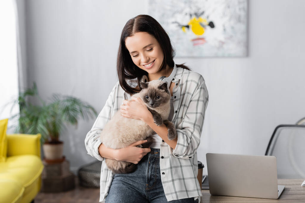 улыбающийся фрилансер, обнимающий кота, стоя дома возле ноутбука - Фото, изображение