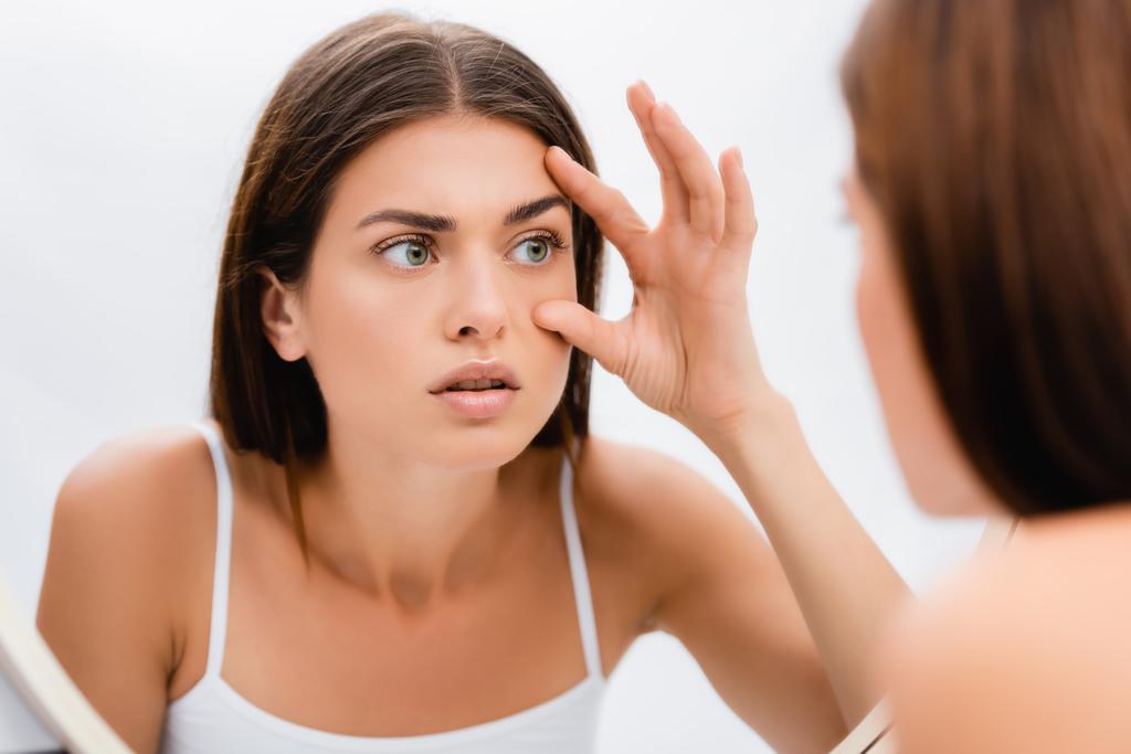 huolestunut nuori nainen katsoo peiliin ja koskettaa kasvoja, hämärtynyt etualalla - Valokuva, kuva