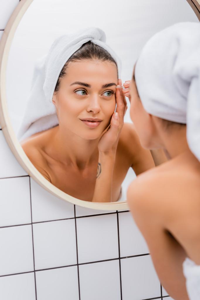 молодая женщина с белым махровым полотенцем на голове выщипывая брови в ванной комнате возле зеркала, размытый передний план - Фото, изображение
