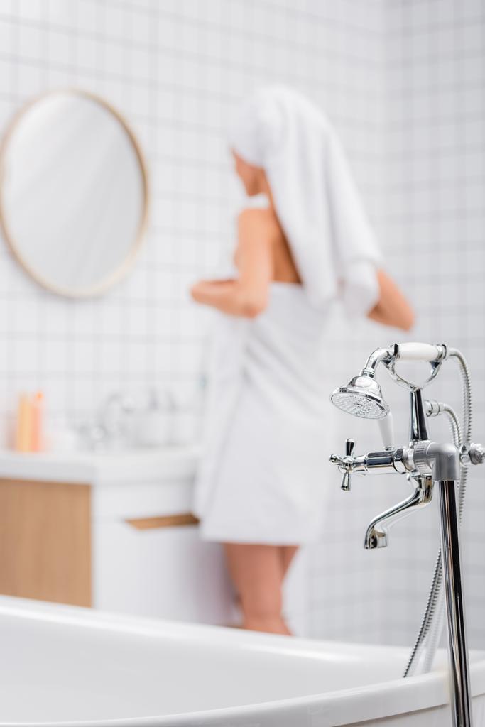 enfoque selectivo de grifo con ducha y mujer envuelta en toallas blancas de pie sobre fondo borroso en el baño - Foto, imagen