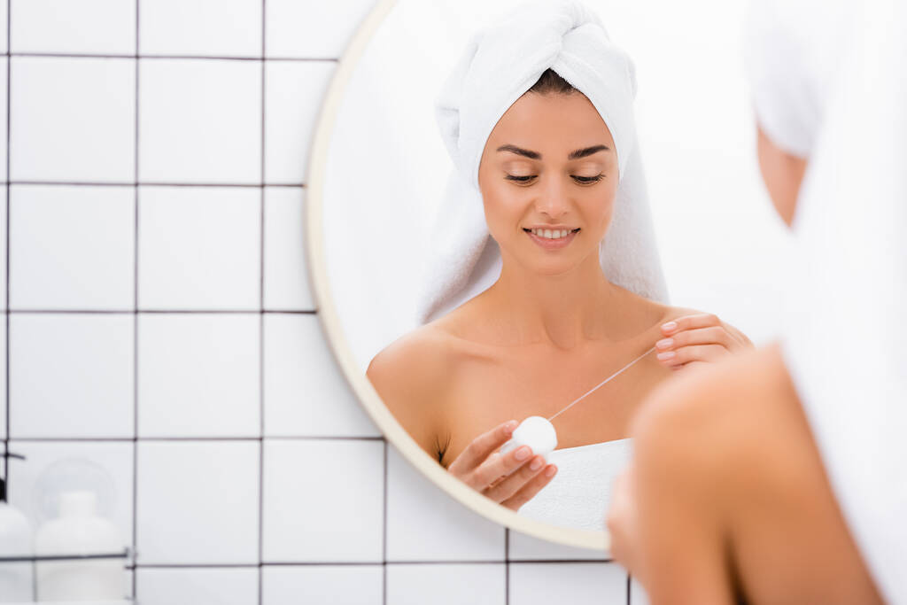 молодая женщина с белым полотенцем на голове держа зубную нить возле зеркала в ванной комнате, размытый передний план - Фото, изображение