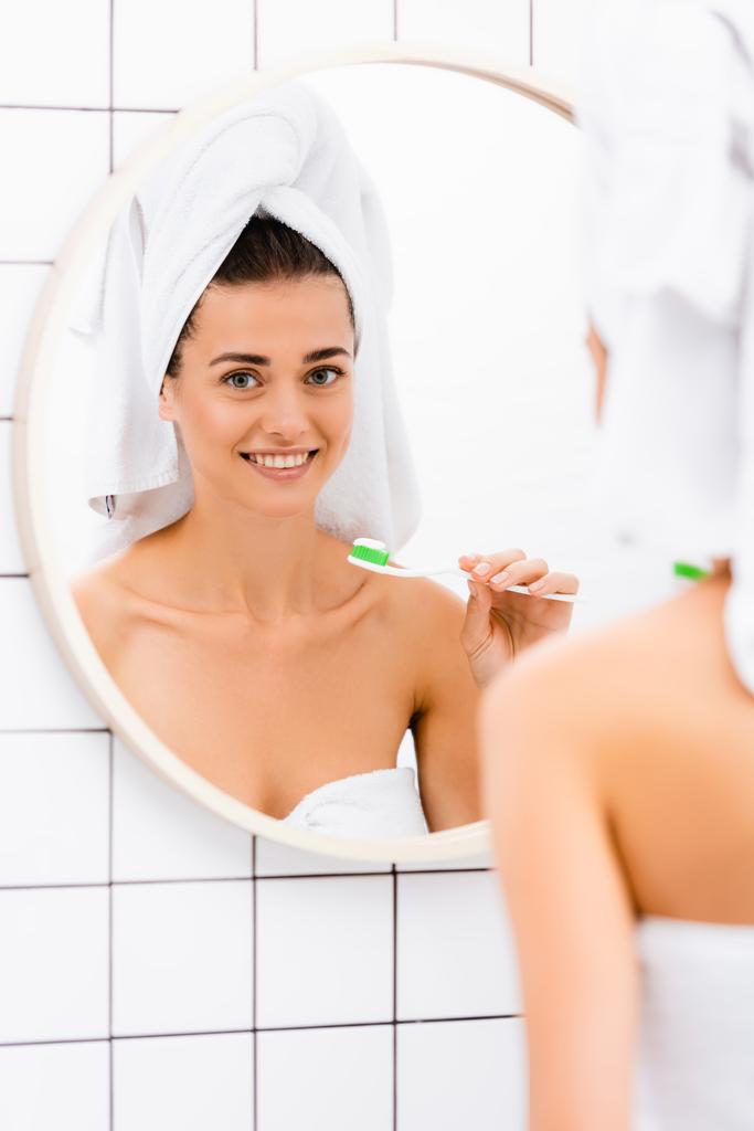 Kafasında beyaz havlu olan mutlu kadın banyoda diş fırçası tutuyor, ön planda bulanıklık var. - Fotoğraf, Görsel