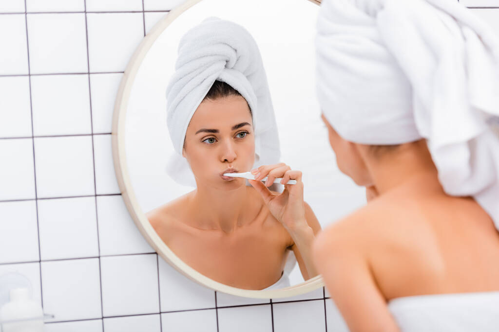 mujer con toalla blanca en la cabeza mirando en el espejo mientras se cepilla los dientes en el baño, borrosa primer plano - Foto, imagen