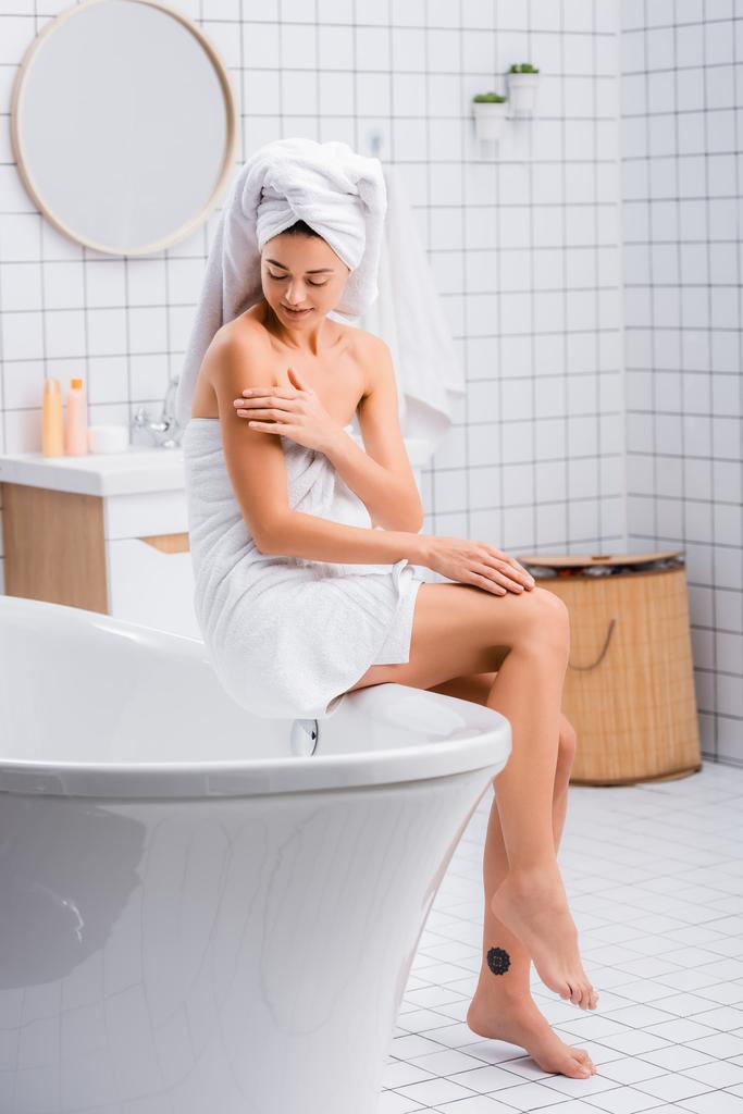 giovane donna, avvolta in asciugamani di spugna bianca, applicando lozione per il corpo mentre seduta sulla vasca da bagno - Foto, immagini