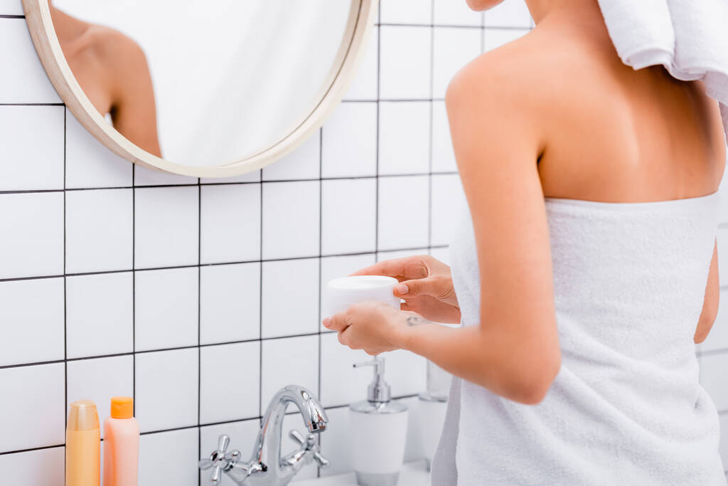 částečný pohled na ženu, zabalenou v bílém ručníku, držící kosmetický krém v koupelně  - Fotografie, Obrázek