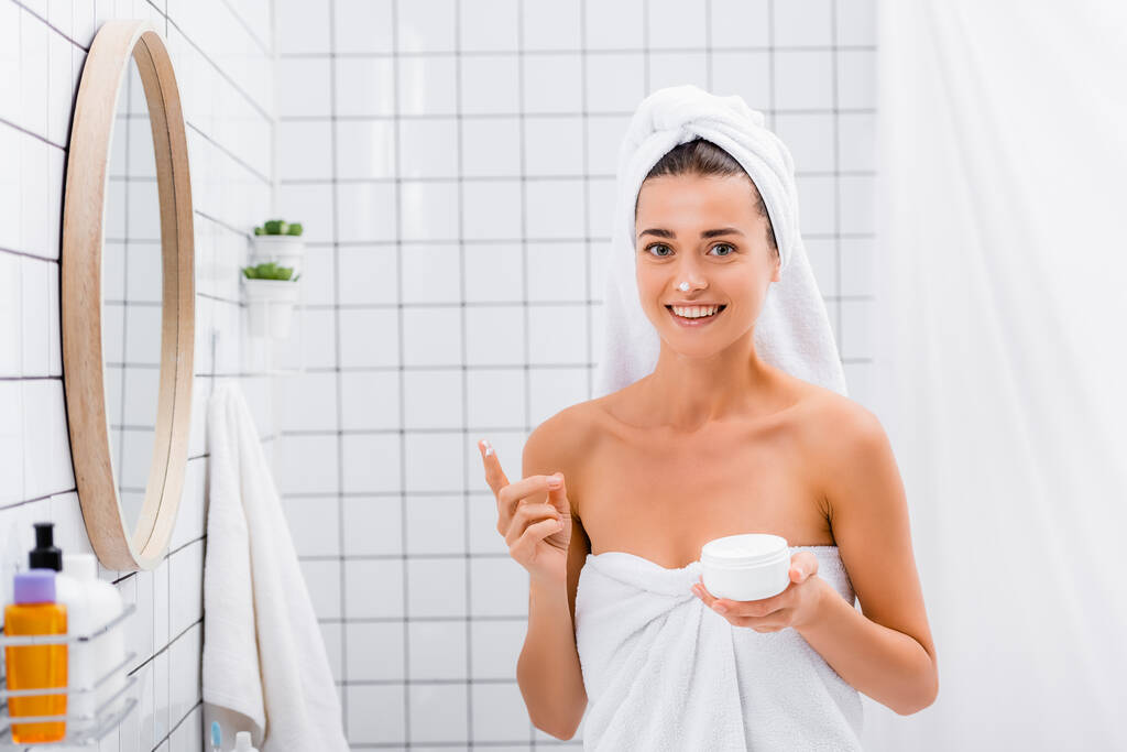 веселая женщина с кремом для лица на носу, улыбающаяся в ванной на камеру - Фото, изображение