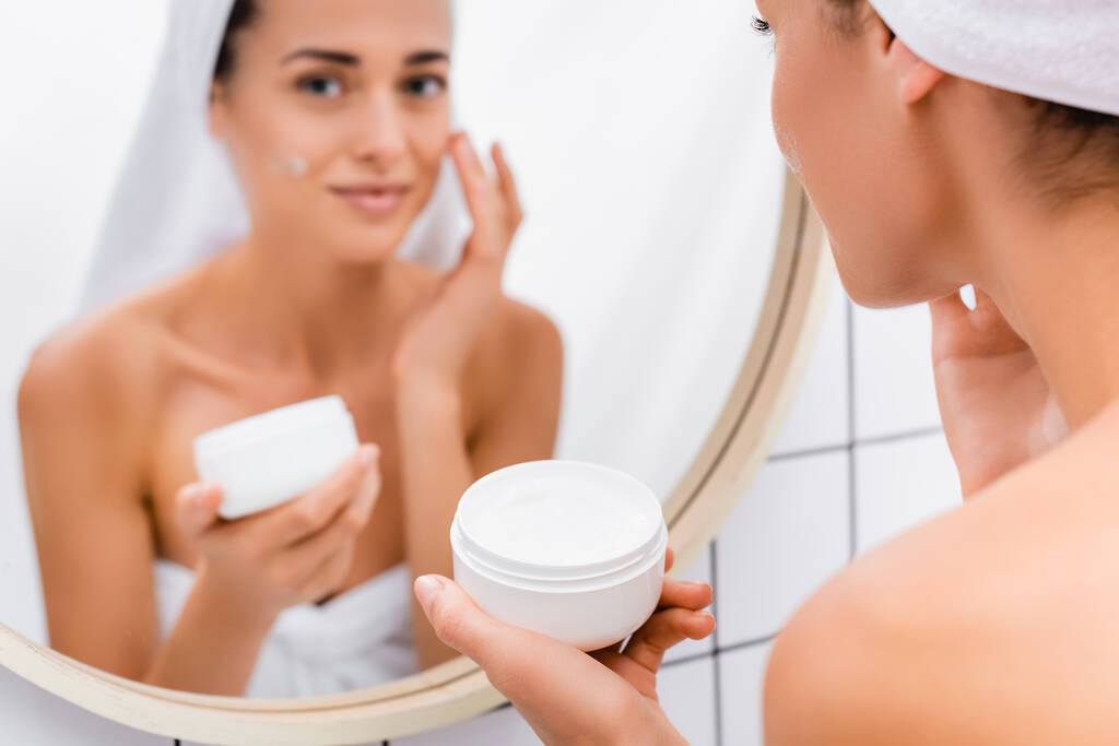 молода жінка наносить крем для обличчя біля розмитого дзеркала у ванній
 - Фото, зображення