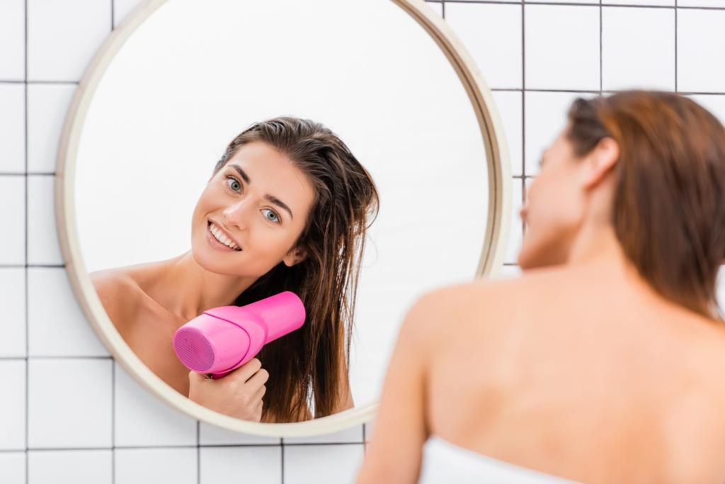 jovem mulher sorrindo enquanto secava o cabelo perto do espelho no banheiro, foreground borrado - Foto, Imagem