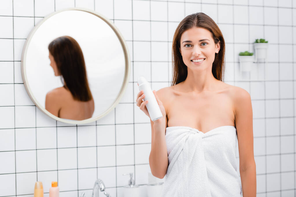 glimlachende vrouw, gewikkeld in witte handdoek, deodorant vasthoudend en glimlachend naar de camera in de badkamer - Foto, afbeelding