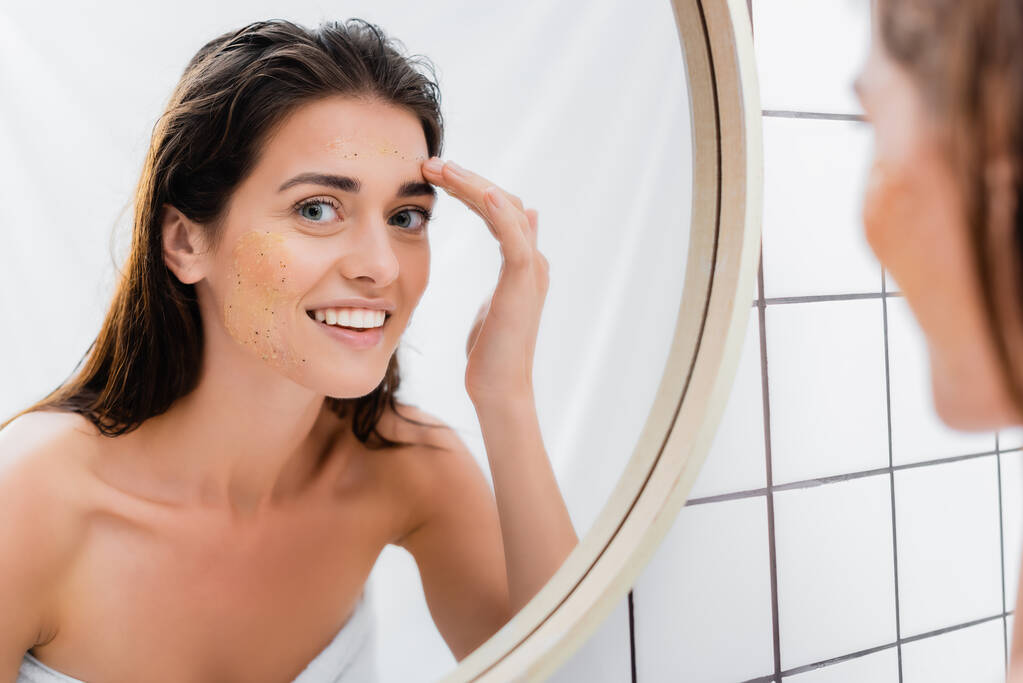 mujer feliz aplicación de exfoliante en la cara cerca del espejo en el baño, borrosa primer plano - Foto, imagen