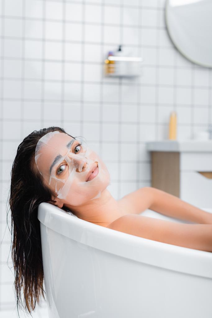 jovem mulher na máscara facial olhando para a câmera enquanto toma banho - Foto, Imagem