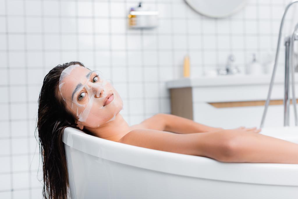 jonge vrouw in het gezicht masker ontspannen in bad en kijken naar de camera - Foto, afbeelding