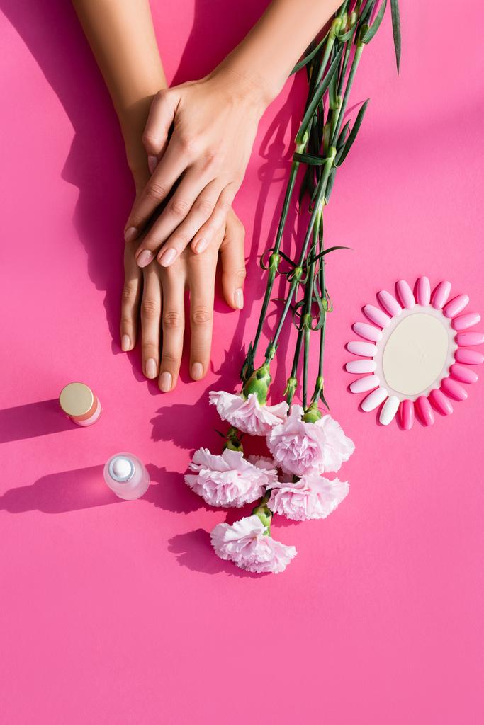 pohled shora na ženské ruce v blízkosti karafiátů, paleta umělých nehtů, odstraňovač kůžičky a lak na nehty na růžovém pozadí - Fotografie, Obrázek