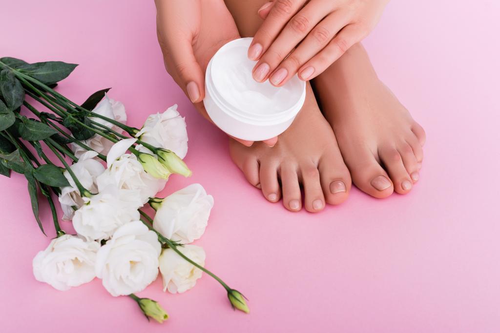 partial view of barefoot woman holding cosmetic cream near white eustoma flowers on pink background - Photo, Image