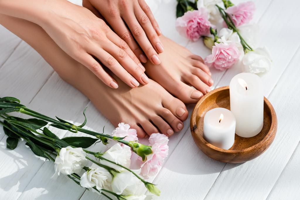 pieds et mains féminins avec des ongles roses brillants près des fleurs d'œillets et d'eustomes et des bougies sur une surface en bois blanc - Photo, image