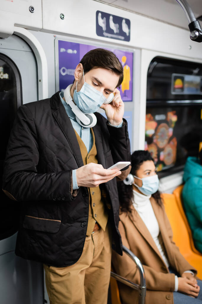 Tıbbi maskeli ve kulaklıklı bir adam metroda Afrikalı Amerikalı bir kadının yanında akıllı telefon kullanıyor.  - Fotoğraf, Görsel
