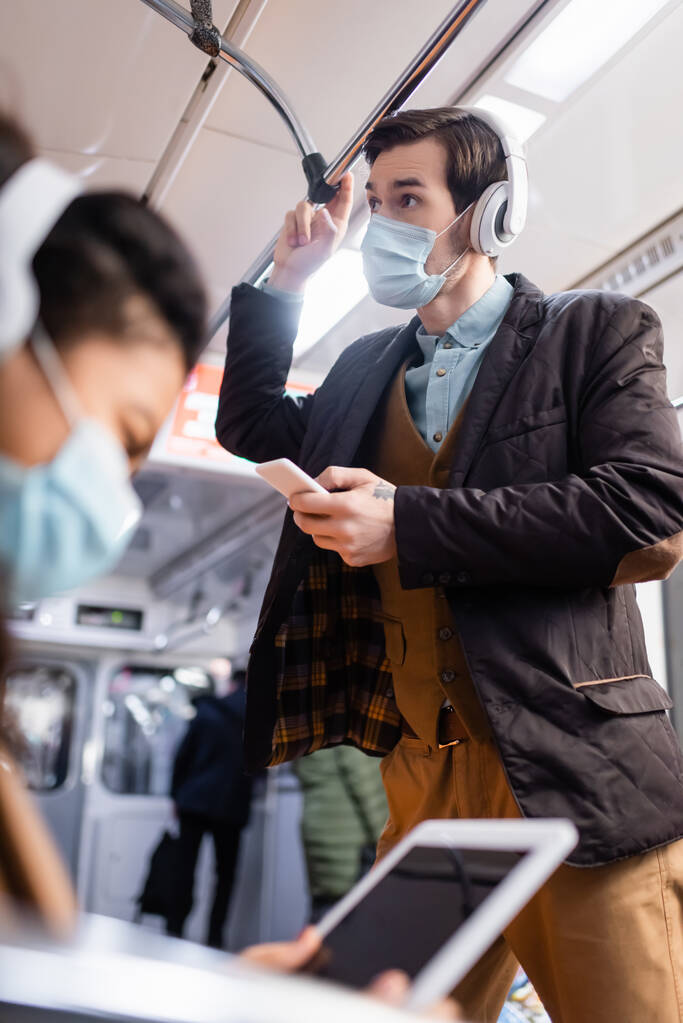 hombre en auriculares con teléfono inteligente cerca de la mujer afroamericana en máscara médica utilizando tableta digital en primer plano borrosa  - Foto, Imagen