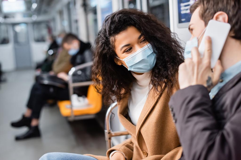 african american woman in medical mask looking at man talking on smartphone on blurred foreground - Photo, Image