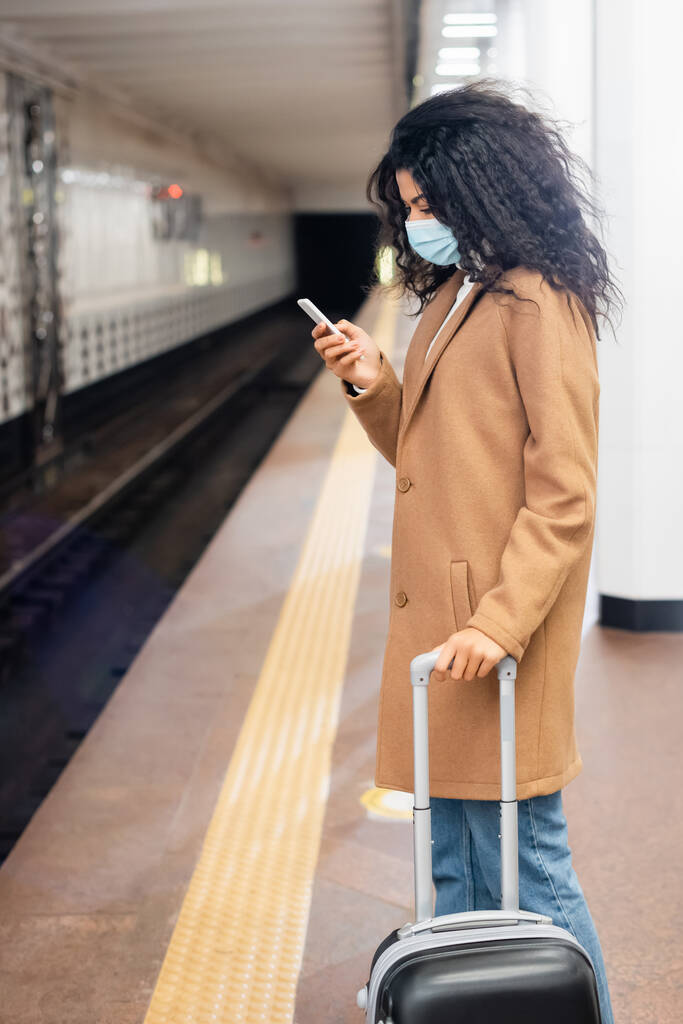 mujer afroamericana en máscara médica de pie con el equipaje y el uso de teléfono inteligente en el metro  - Foto, Imagen