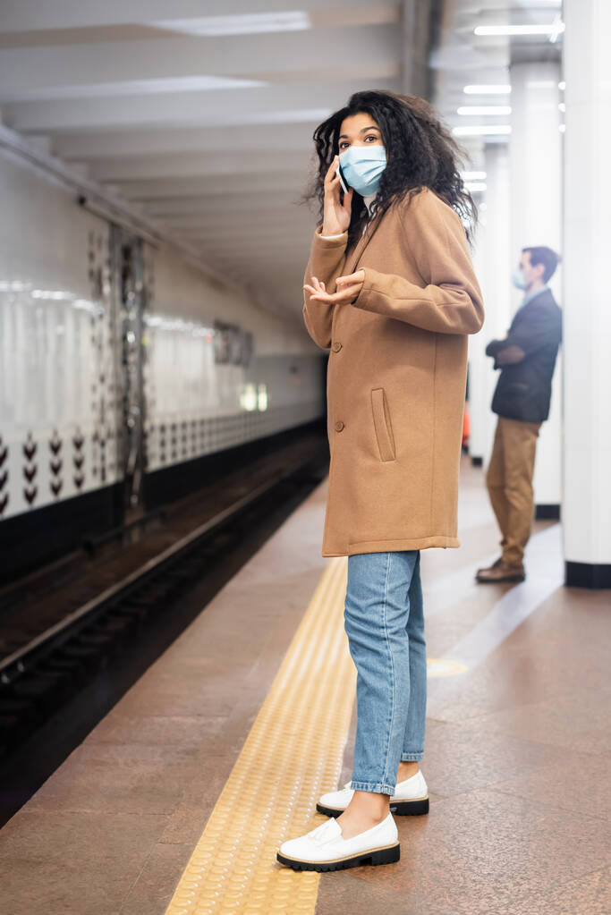 pleine longueur de jeune femme afro-américaine en masque médical parlant sur smartphone dans le métro - Photo, image