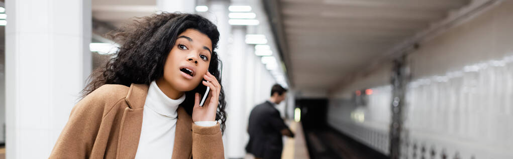 Şaşırmış Afrikalı Amerikalı kadın akıllı telefondan konuşuyor ve bulanık arka planda metroya bakıyor. - Fotoğraf, Görsel