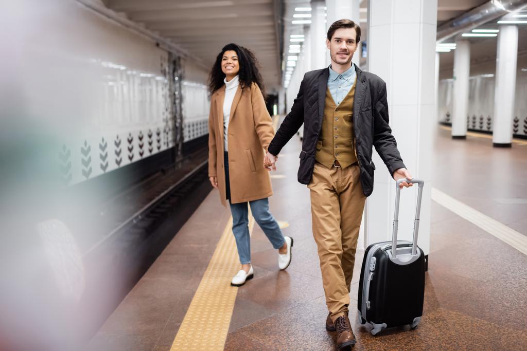interrazziale coppia che si tiene per mano in metropolitana mentre si cammina con i bagagli su sfondo sfocato - Foto, immagini