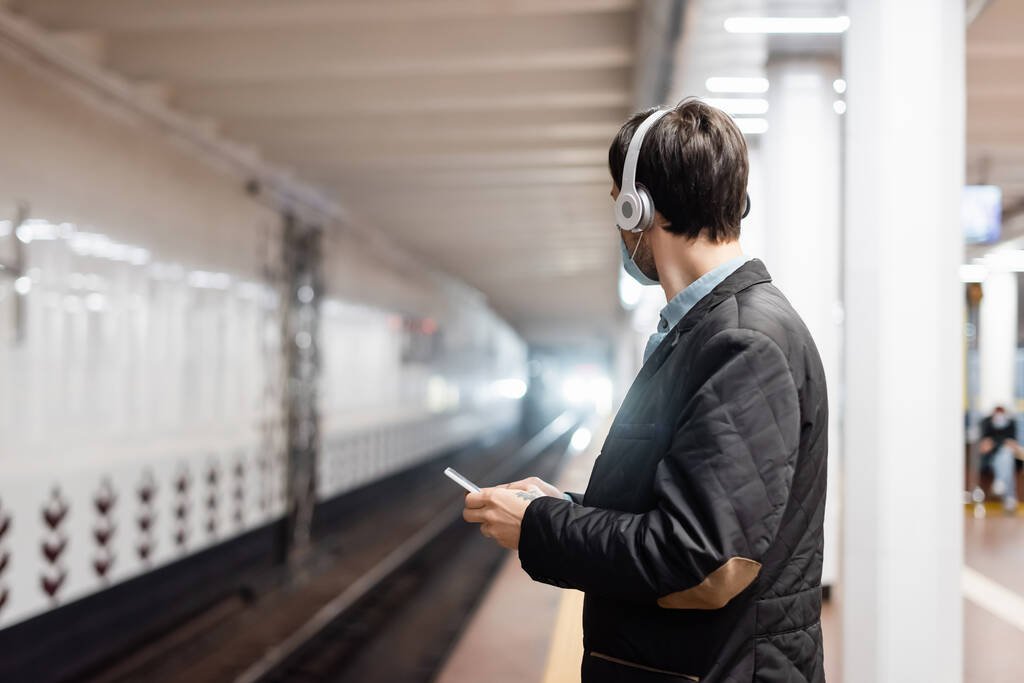 wytatuowany mężczyzna w masce medycznej i słuchawkach bezprzewodowych trzymających smartfona w metrze  - Zdjęcie, obraz