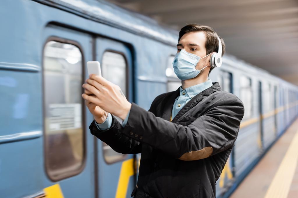 uomo in maschera medica e cuffie senza fili scattare selfie su smartphone in metropolitana  - Foto, immagini